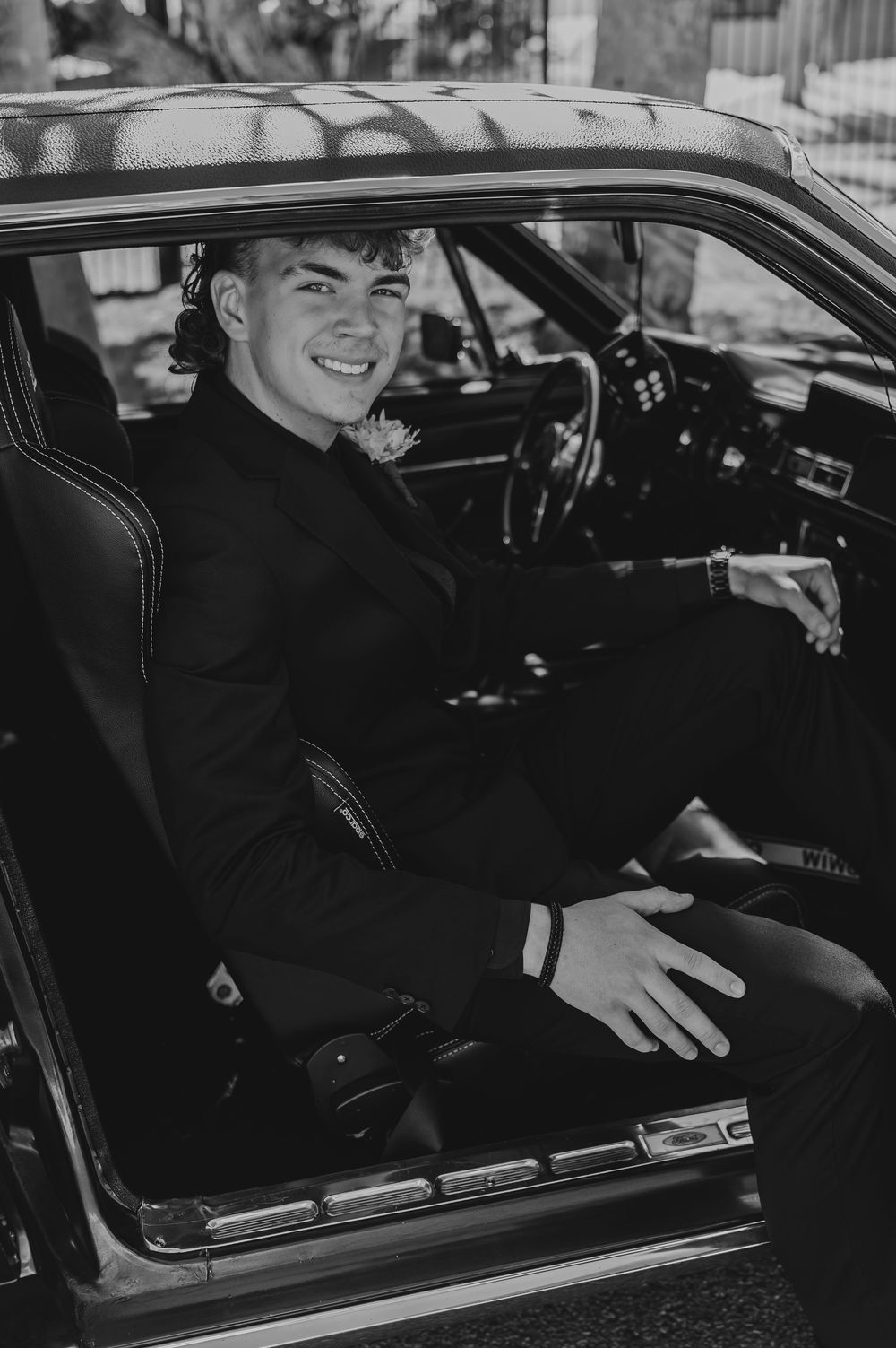 A black and white photo showing someone seated in the driver's seat of a classic car, smiling through the window.