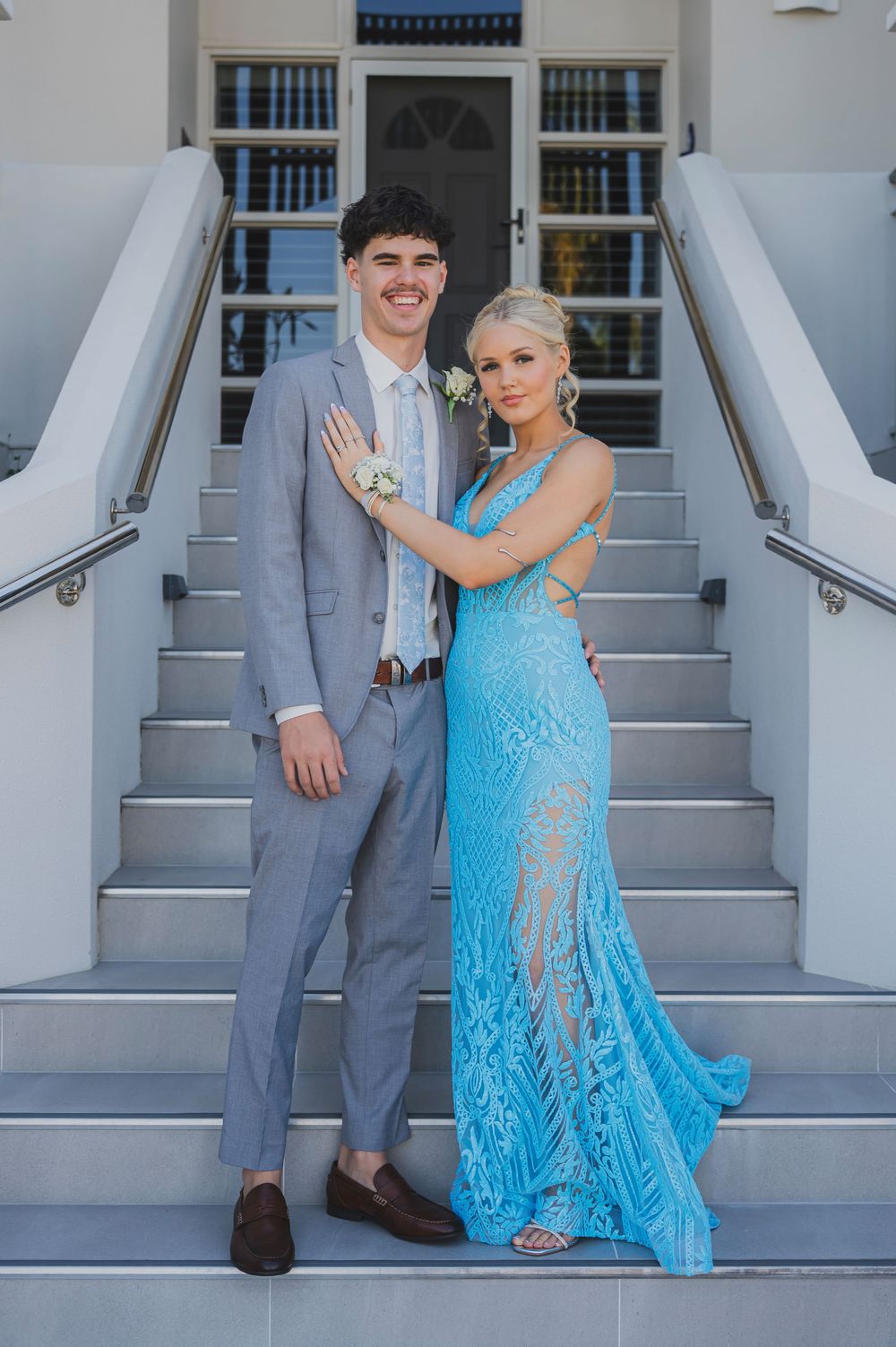 A couple poses playfully on white stairs, with the woman wearing a bright blue lace dress and the man in a gray suit.