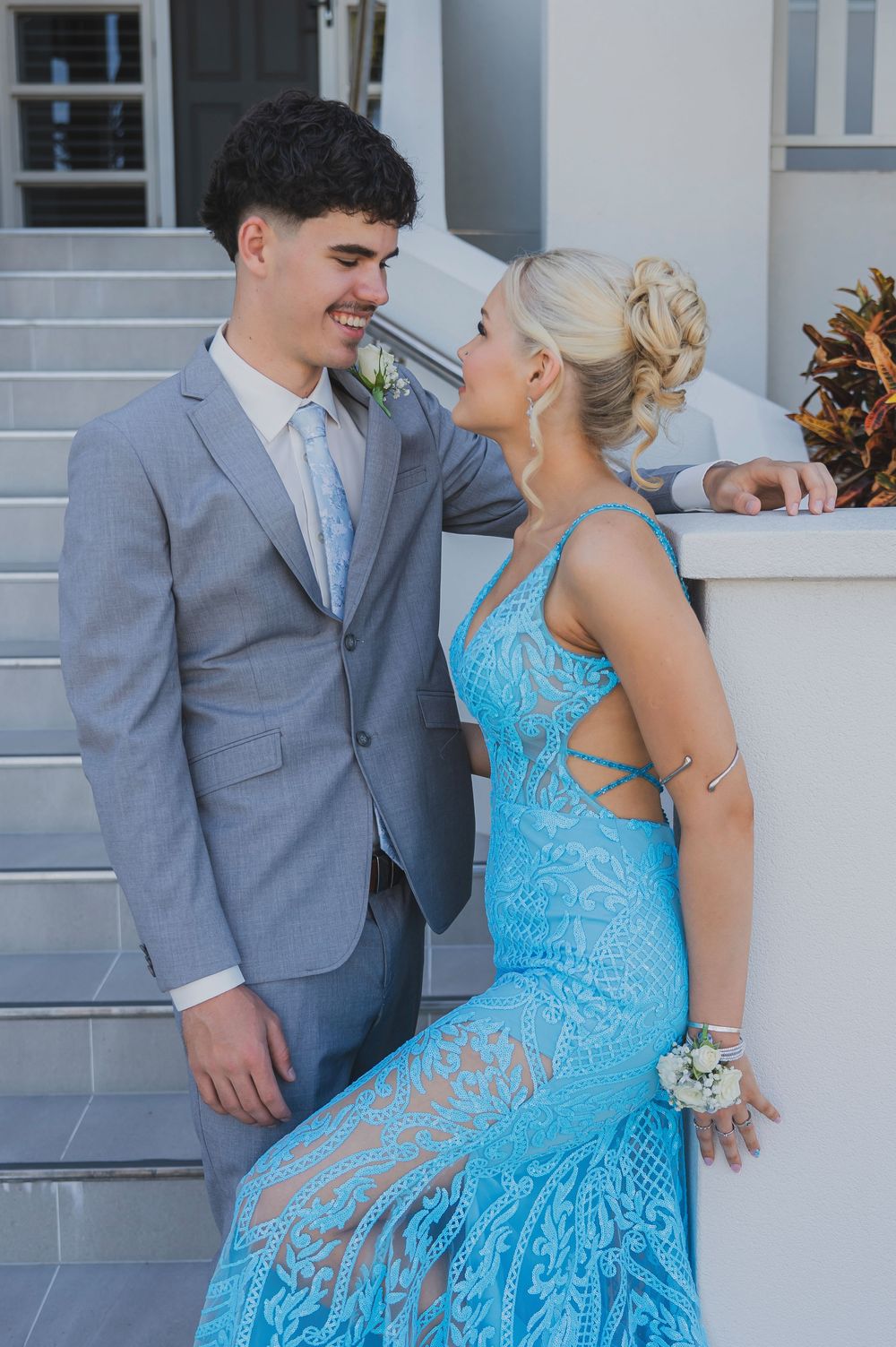A couple in formal attire sharing romantic moments on white stairs, with her wearing a turquoise dress and him in grey.