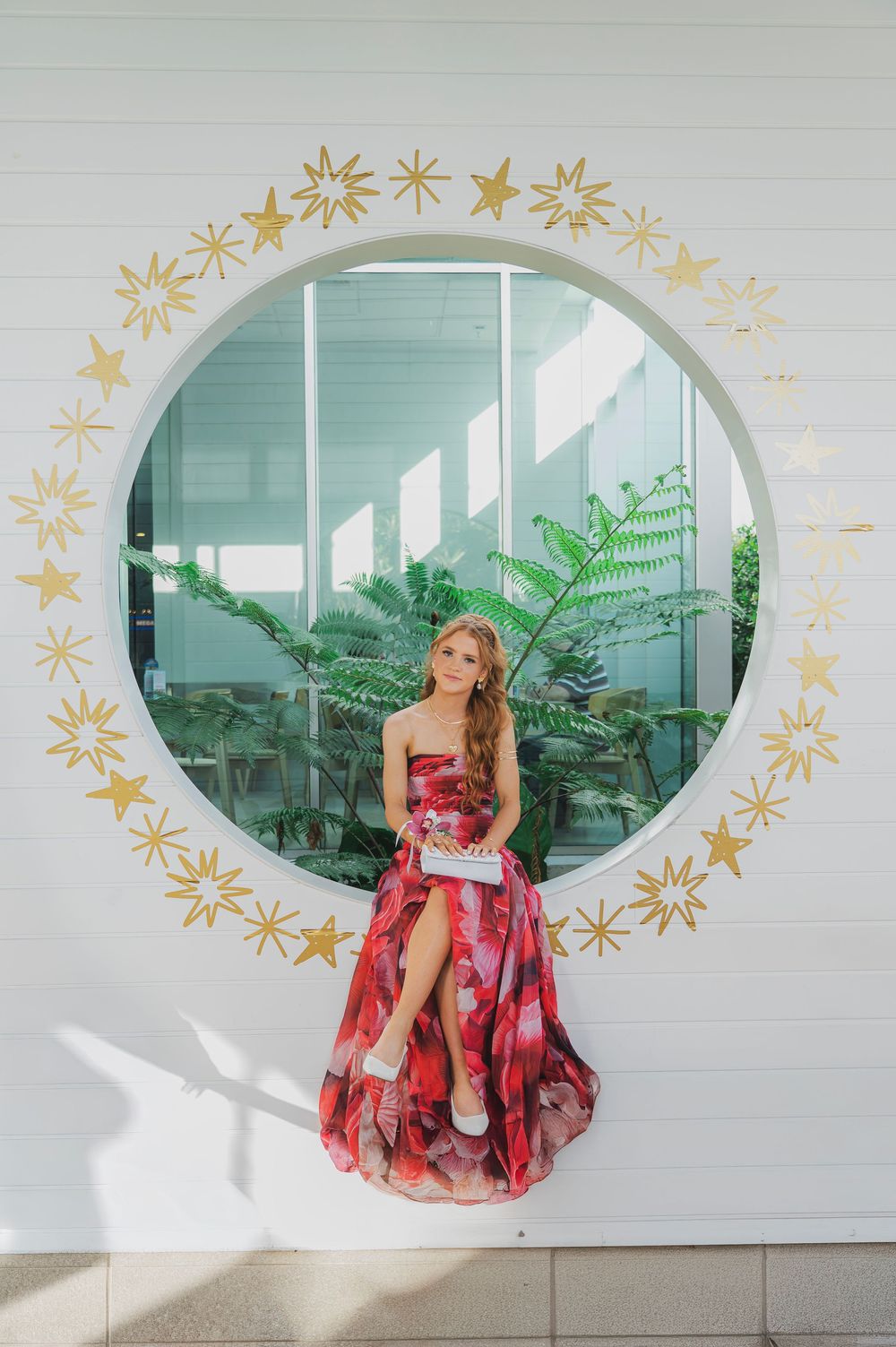 A person in a flowing red dress poses in front of a white wall with decorative gold stars and an oval window.