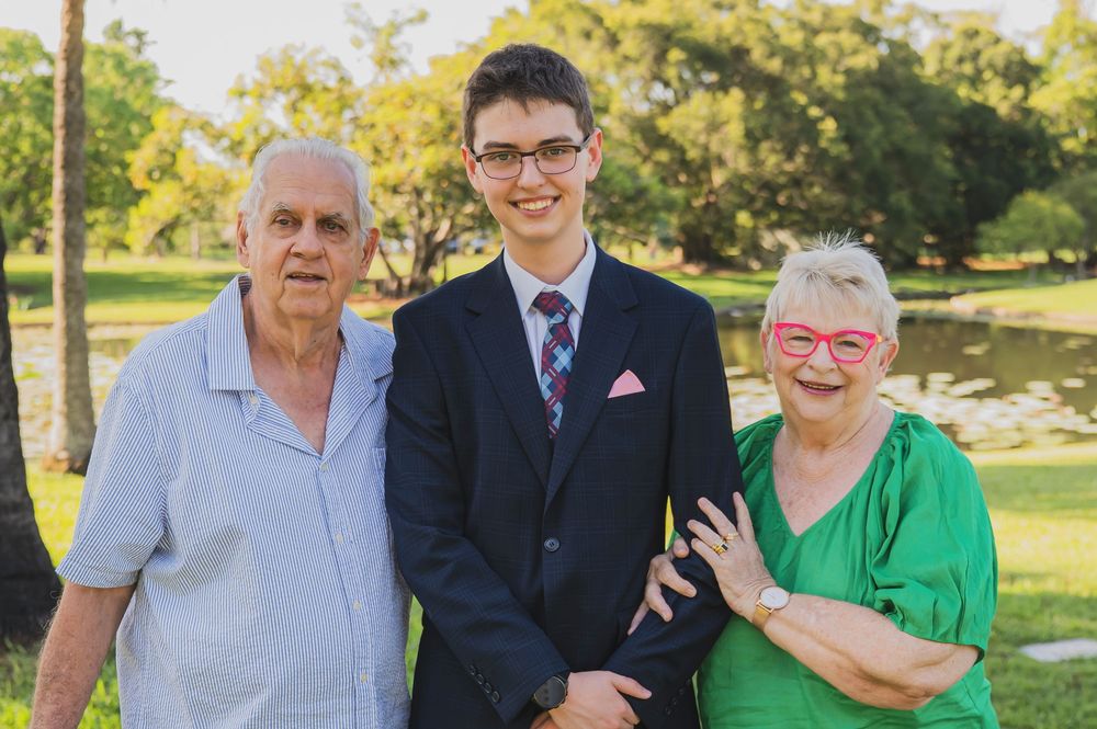 A group of people in formal and casual attire stand together outdoors in a sunny park setting.