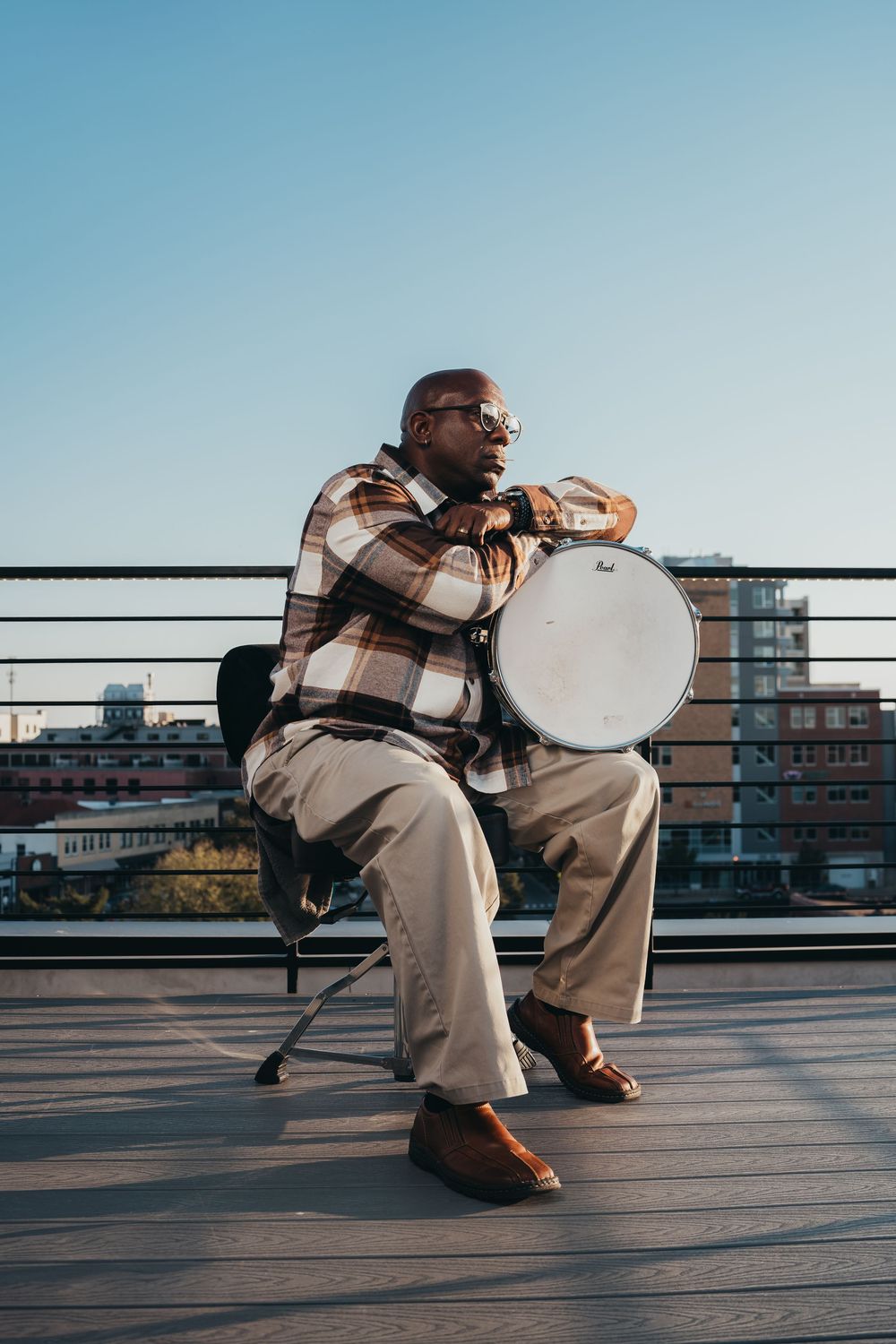 Musician playing drum while sitting on rooftop with urban skyline backdrop.