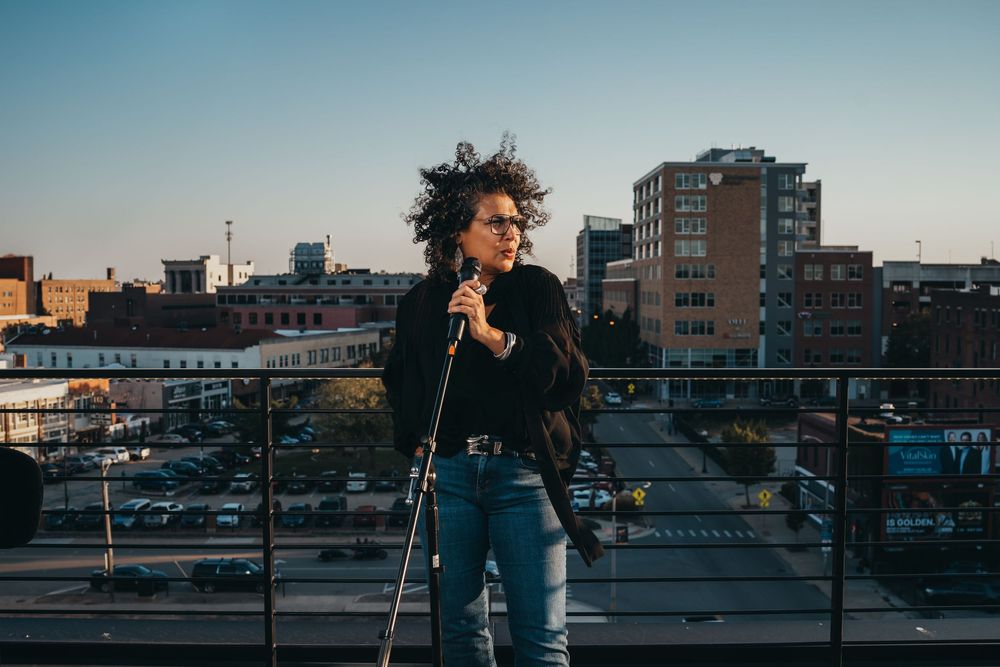 Performer singing on a rooftop with city buildings in the background during sunset.