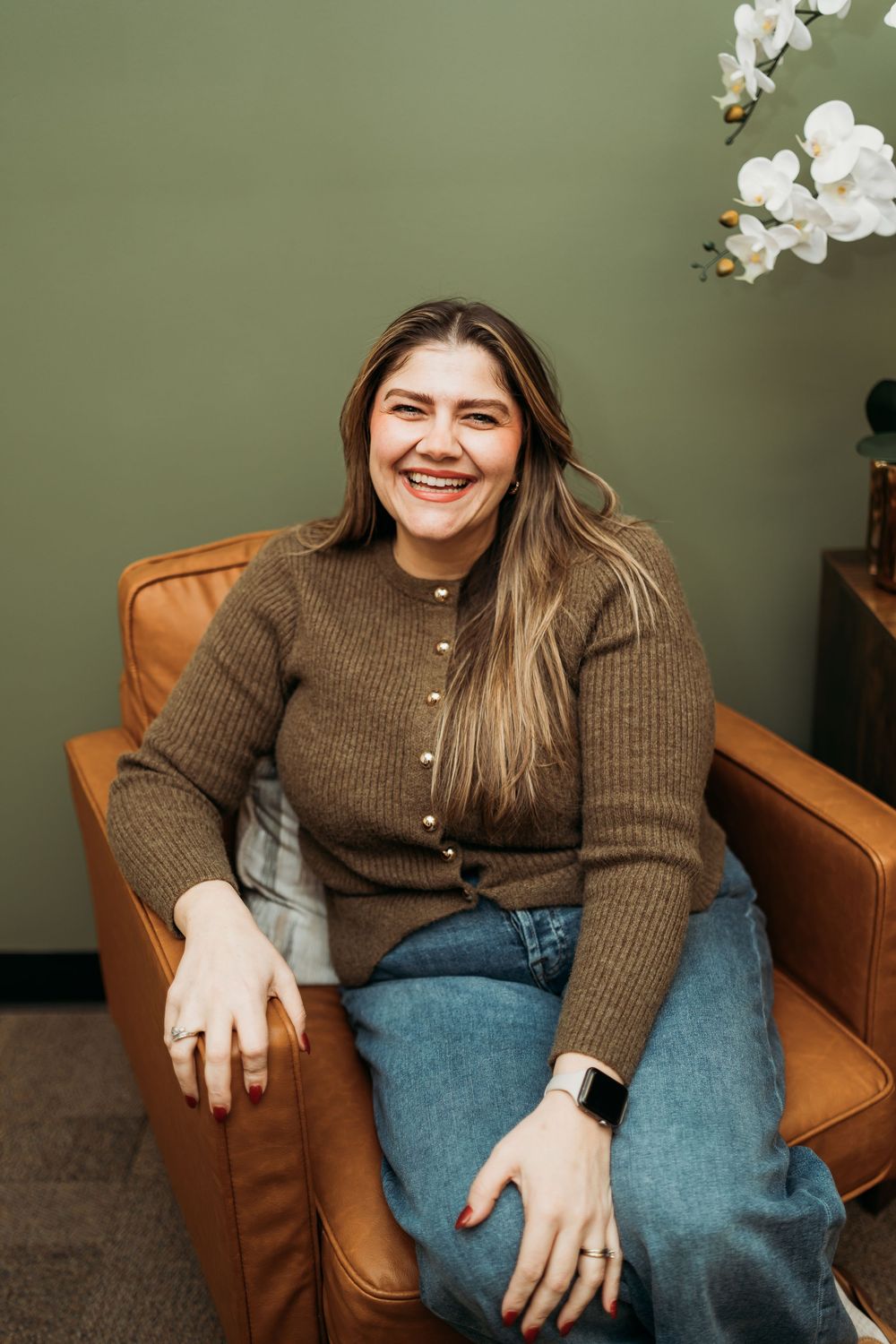 Person in brown sweater and jeans relaxing in leather chair against green wall.