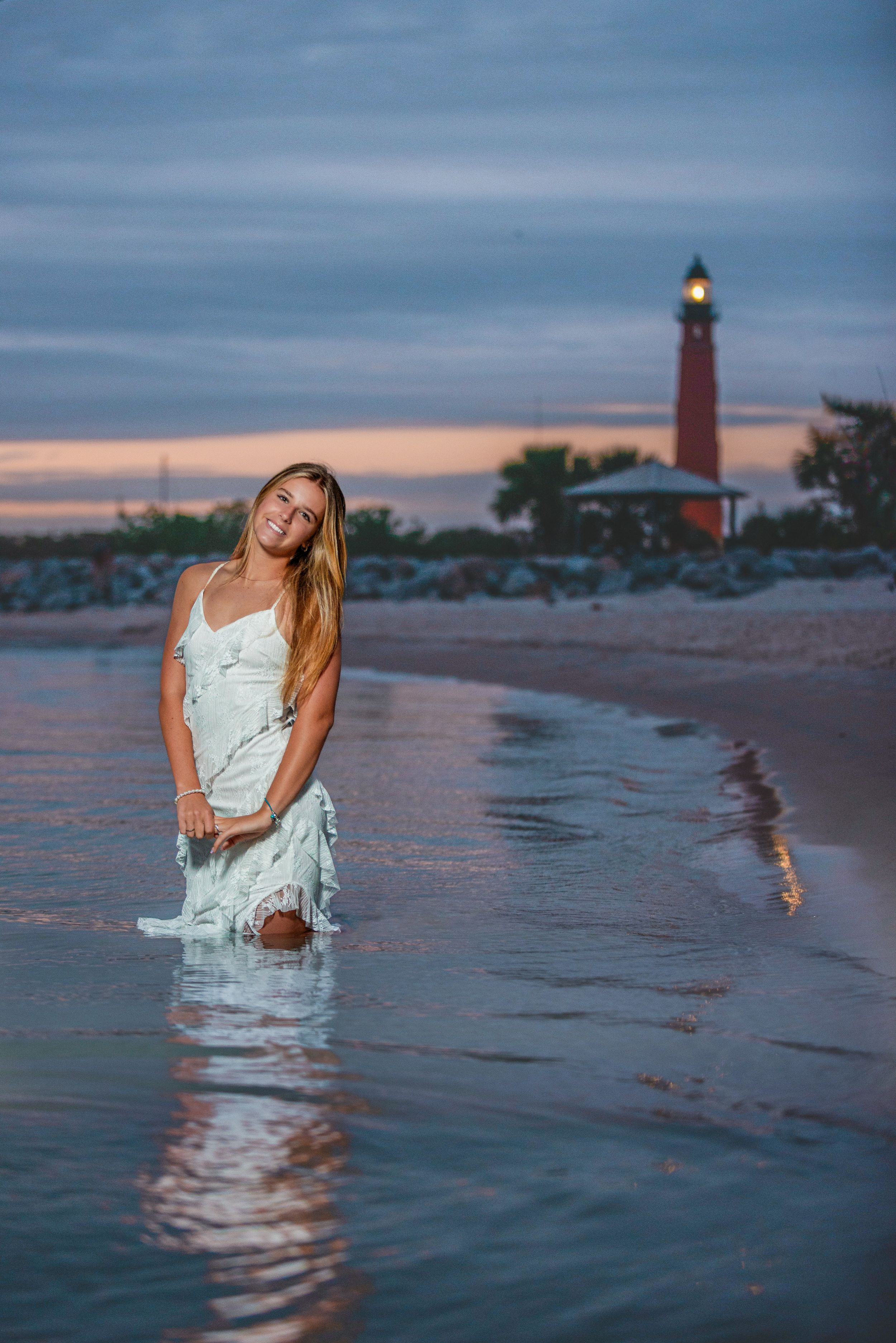 Young woman in white dress standing in ocean waves at sunset with a red lighthouse glowing in the background.