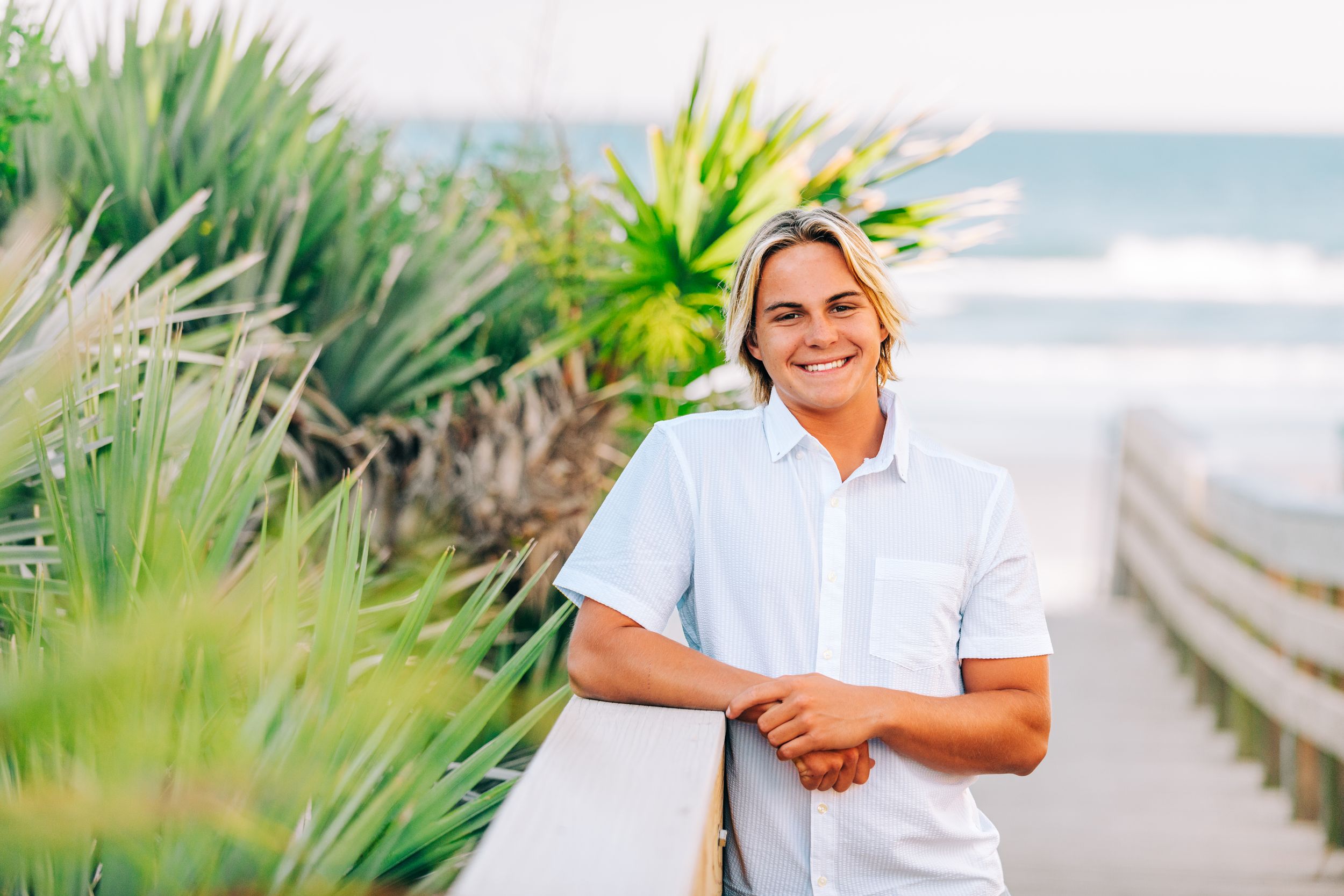 Smiling young man with blonde hair in white shirt leaning on railing by tropical plants and ocean beach.
