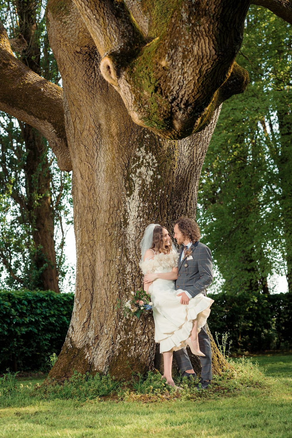 A romantic wedding couple embraces beneath a majestic old tree in a lush green garden setting.