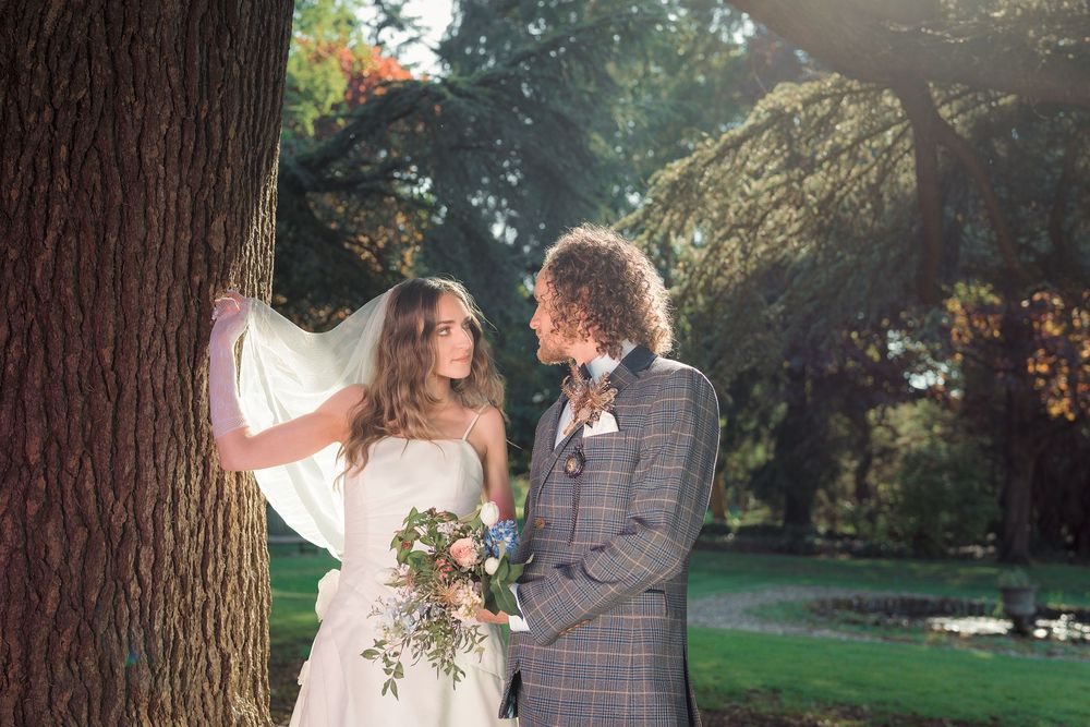 Couple in wedding attire embracing under a large tree in a scenic outdoor garden setting.
