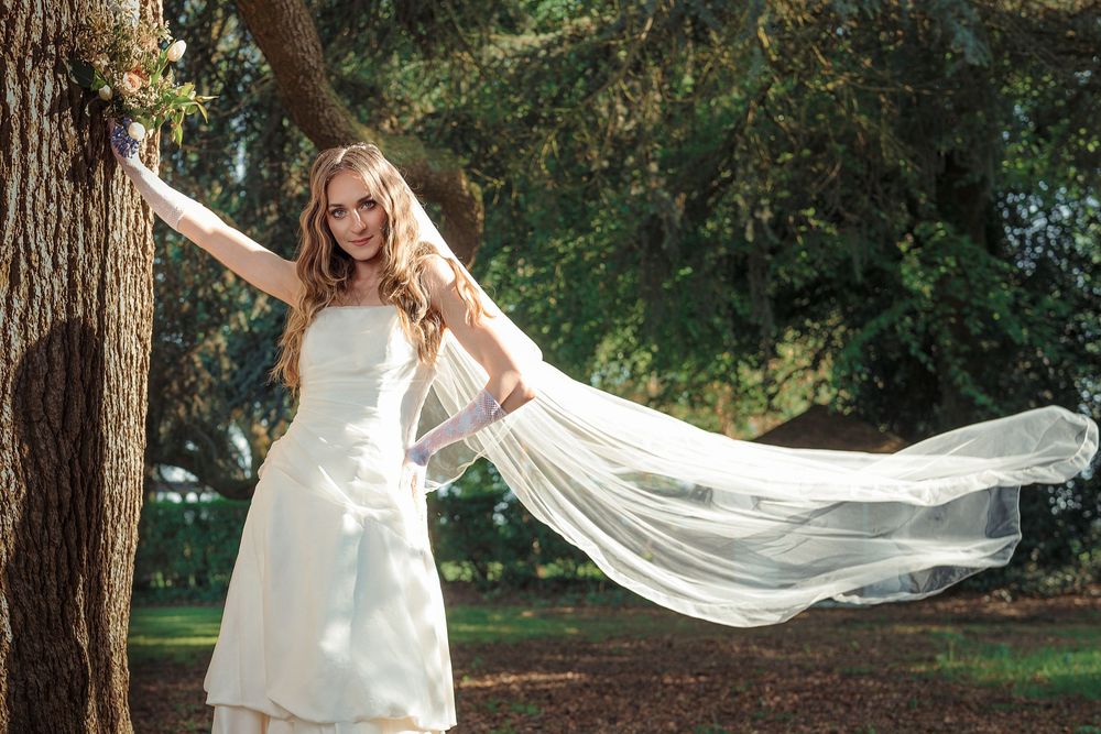 A flowing white veil catches the breeze as a bride poses among tall trees in a forest setting.