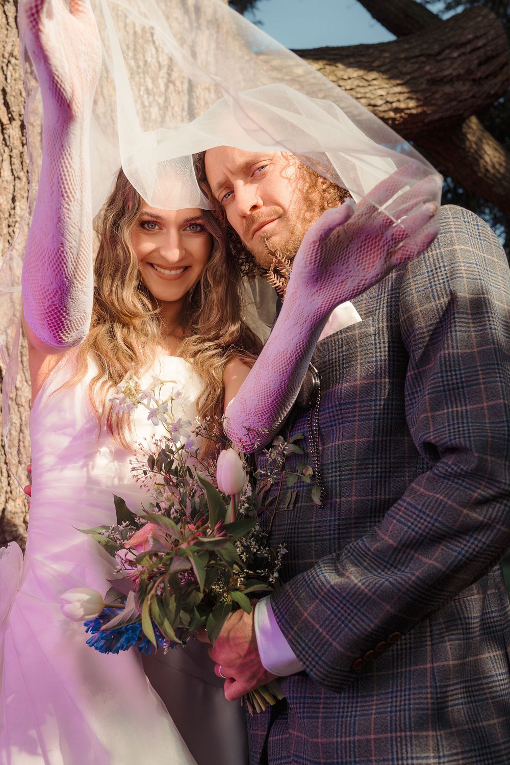A romantic wedding couple embraces under a flowing veil with sunlight creating a dreamy pink glow in the background.