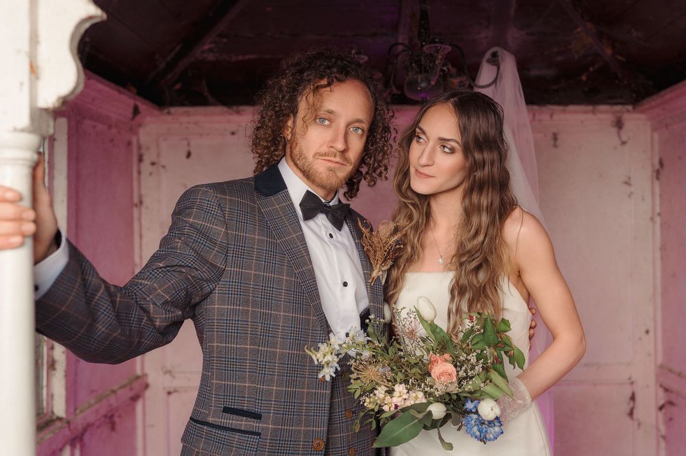 A couple in formal wedding attire pose together in a pink room with distressed walls.