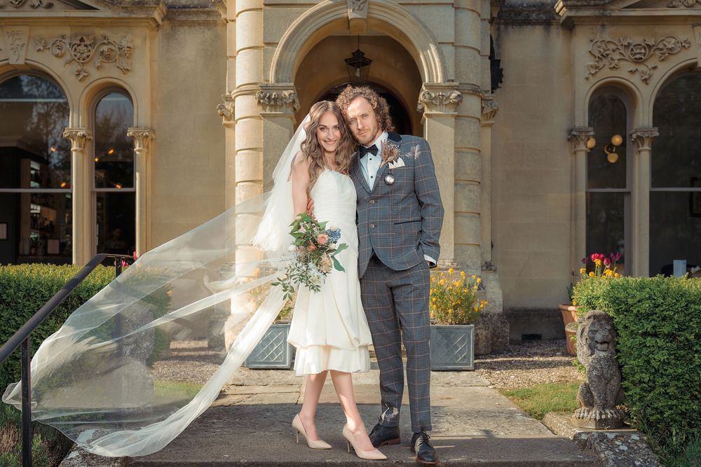 Wedding couple poses together at elegant stone venue with arched windows and flowing veil in the breeze.