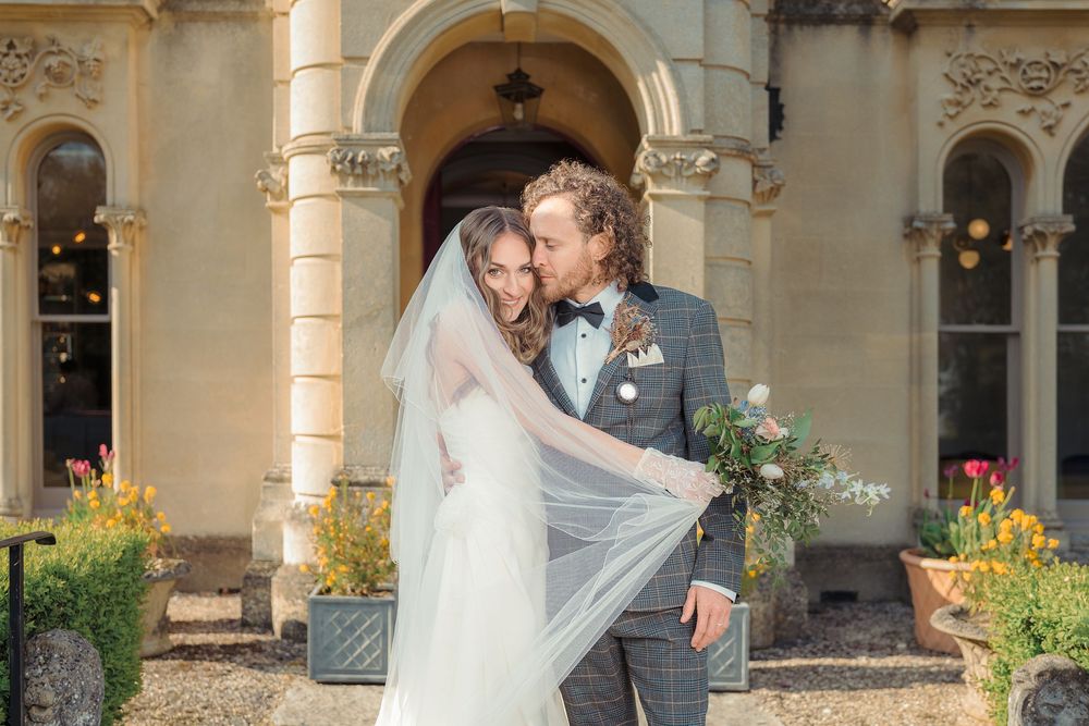 A couple shares a romantic moment on their wedding day in front of an elegant stone archway with blooming flowers.