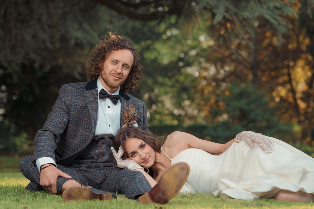 Wedding couple relaxes on grass in autumn setting wearing formal attire.