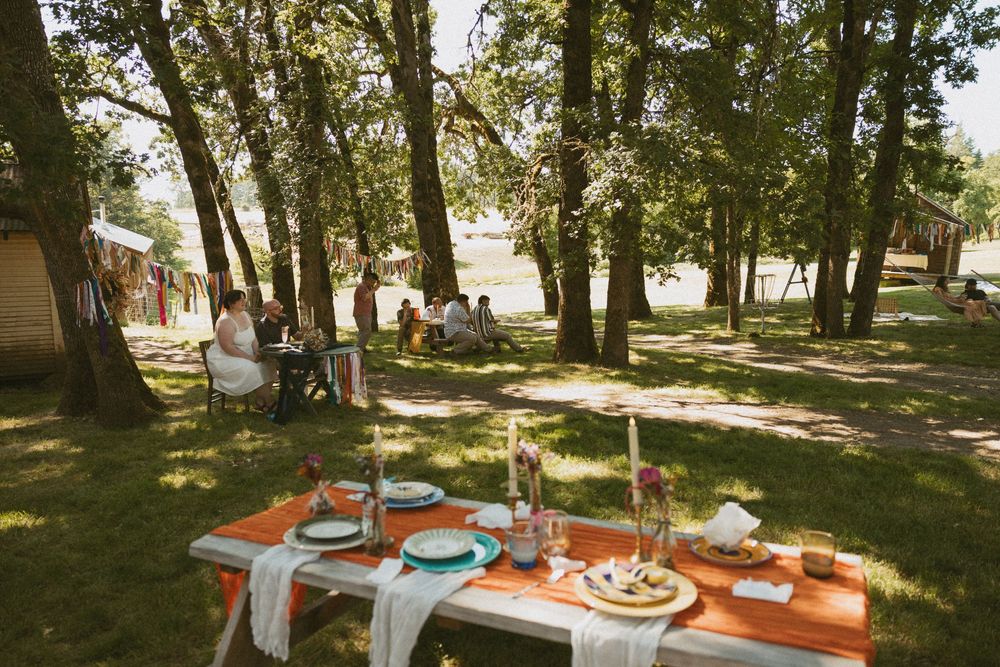Outdoor picnic table set for a meal with colorful dishes and glasses under shady trees in a park-like setting.