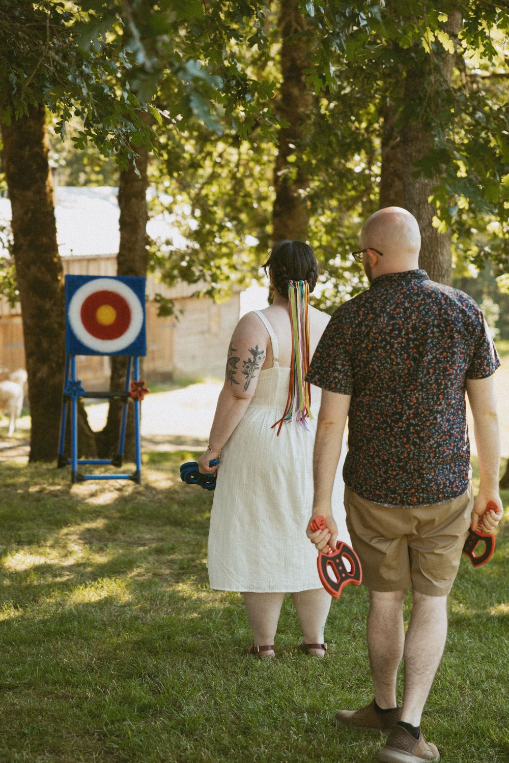 Archers stand in a grassy outdoor area with archery targets visible in the background during a sunny summer day.