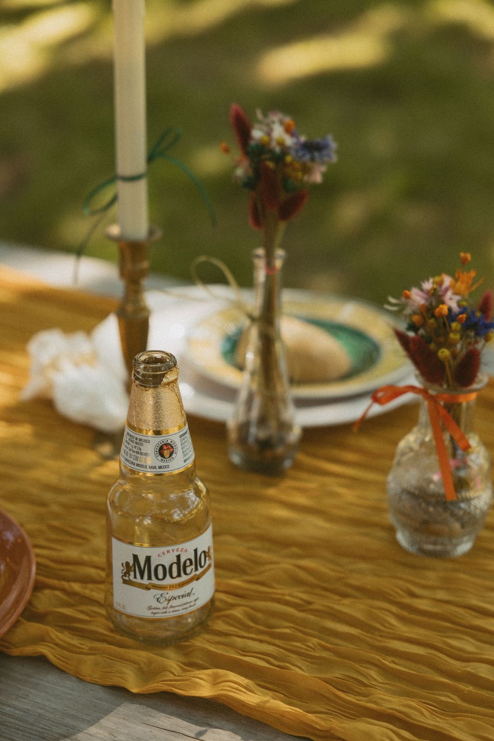 Small glass vases with wildflowers and a Modelo beer bottle arranged on a golden tablecloth for rustic table decor.