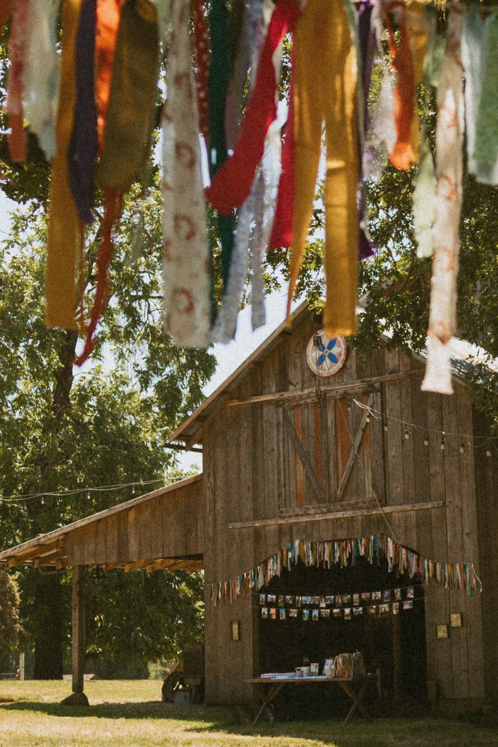 Rustic wooden barn decorated with colorful hanging ribbons and a star emblem under tall trees in a rural setting.