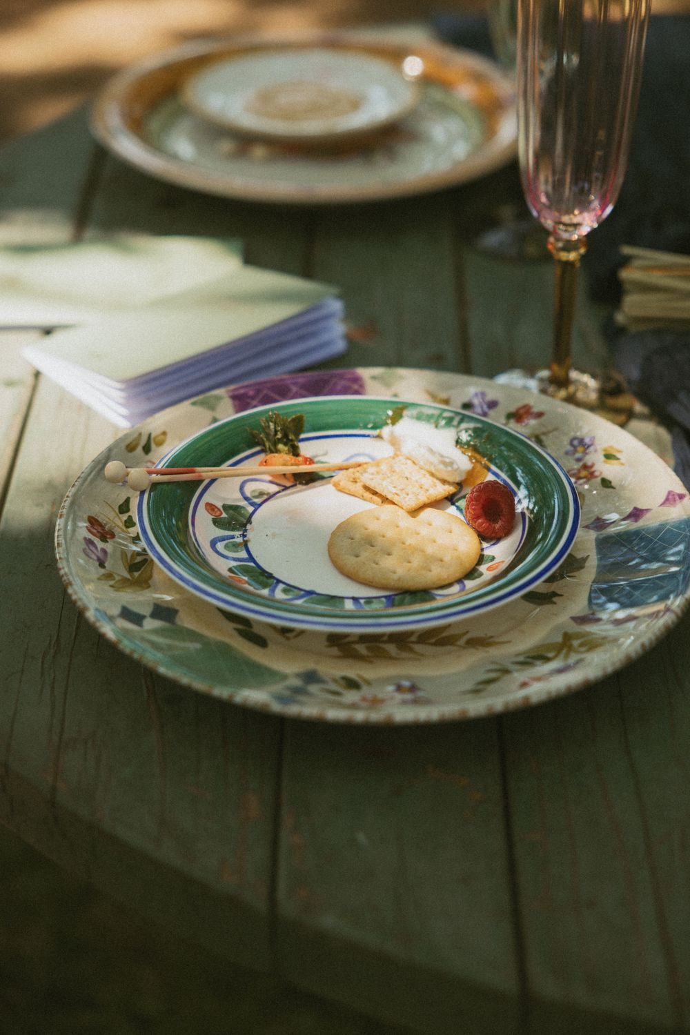 A decorative plate with crackers and cheese sits on a green wooden table alongside a wine glass and napkins.