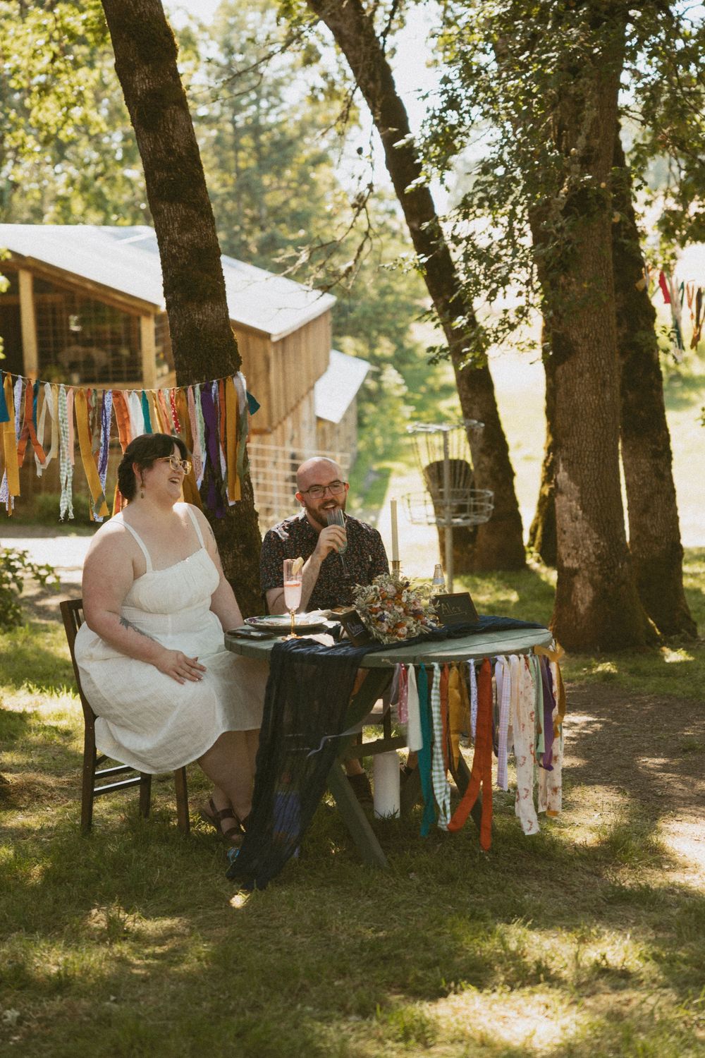 A couple sits at a rustic outdoor table decorated with colorful fabric streamers during a woodland celebration.