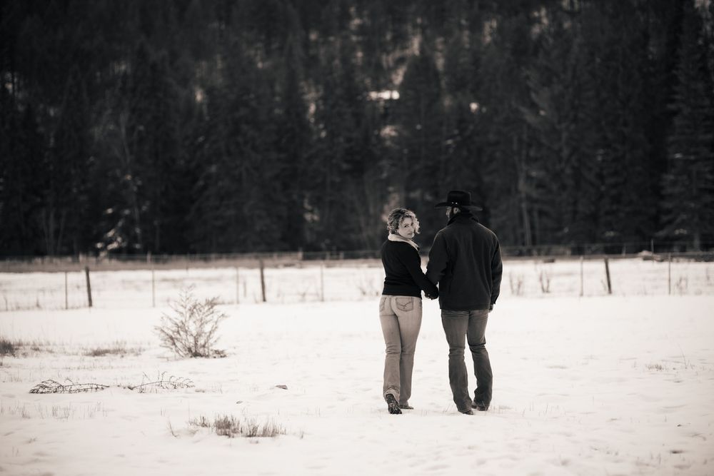 Two people walking together across a snow-covered field with evergreen forest in the distance.