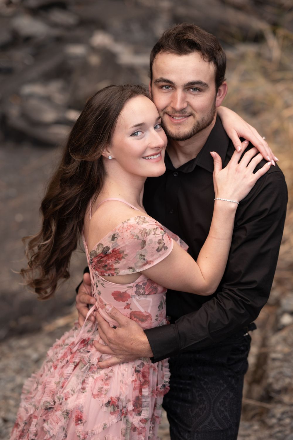 Romantic couple embracing outdoors, woman wearing floral pink dress against natural backdrop.