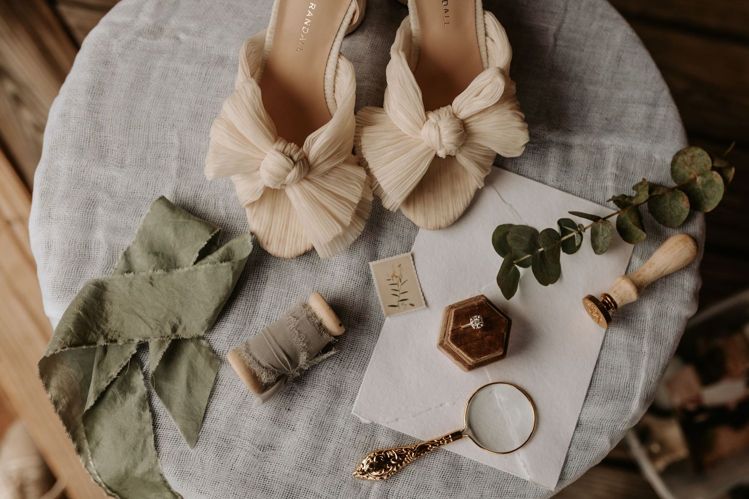 Wedding accessories including beige bow heels, eucalyptus, rings, and decor arranged on gray linen.