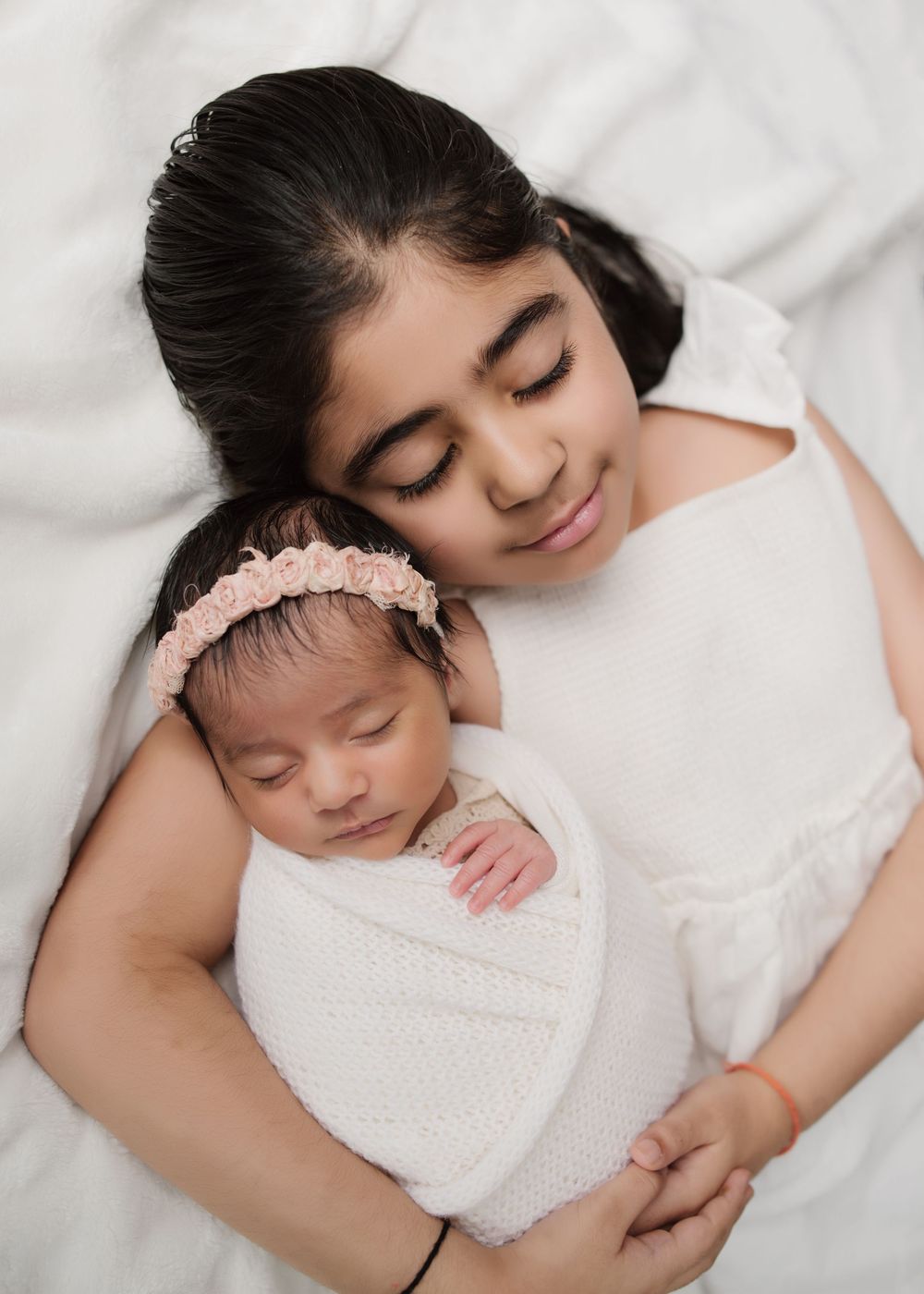 Sibling sleeping peacefully in white wrap and pink headband during an in-home newborn photography session