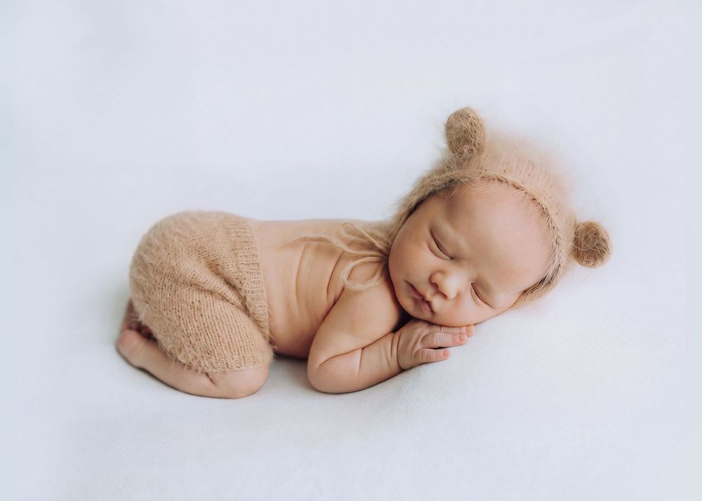 Newborn baby boy sleeping peacefully in a soft beige knitted bear bonnet and matching shorts during an in-home newborn photography session