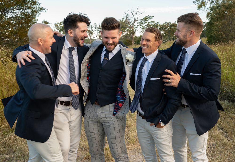 Groom laughing with four groomsmen in navy suits in rustic outdoor Australian bush setting.