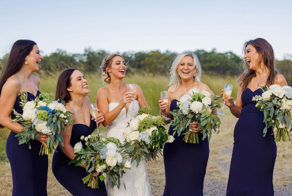 Bride laughing with four bridesmaids in navy gowns holding white bouquets in sunny field.