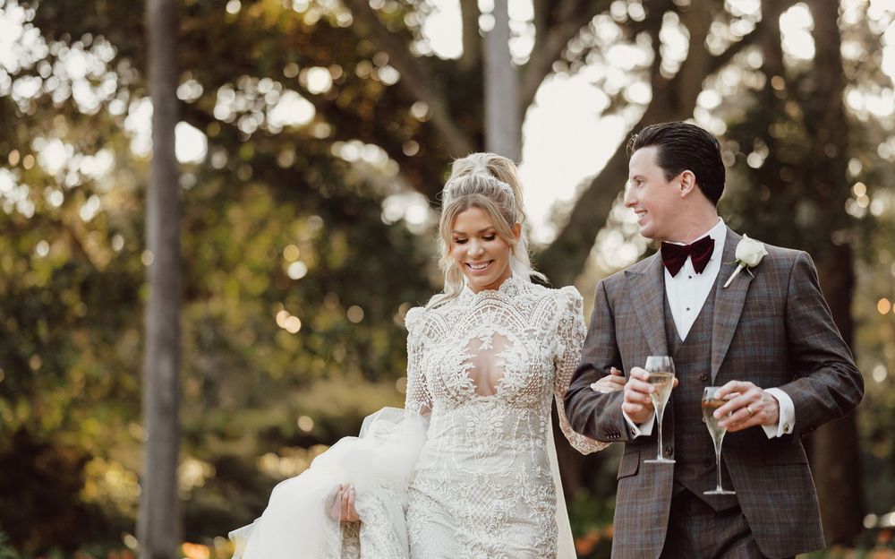 Elegant bride in lace gown and groom in grey suit walk together holding champagne glasses outdoors.