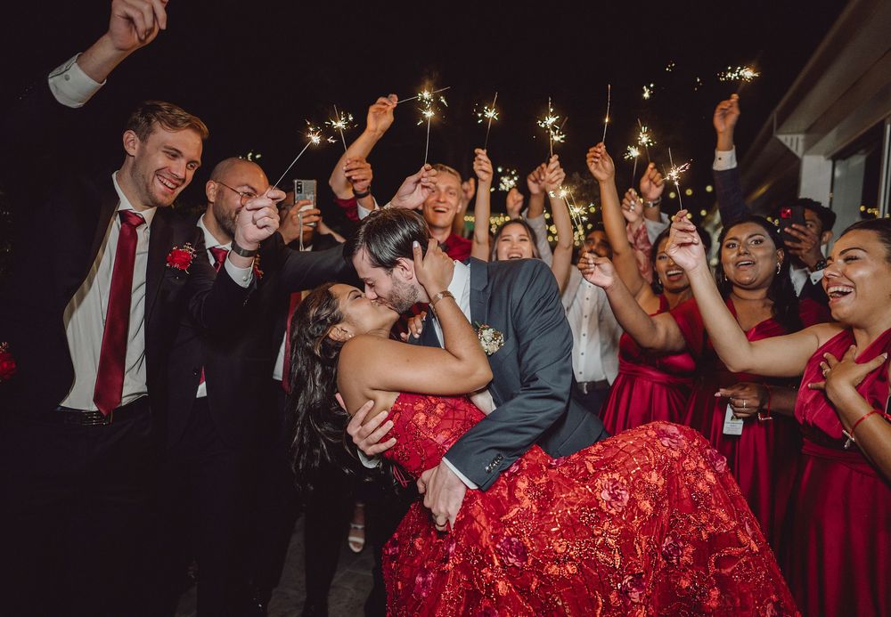 Couple shares romantic kiss surrounded by cheering guests holding sparklers at night wedding celebration.
