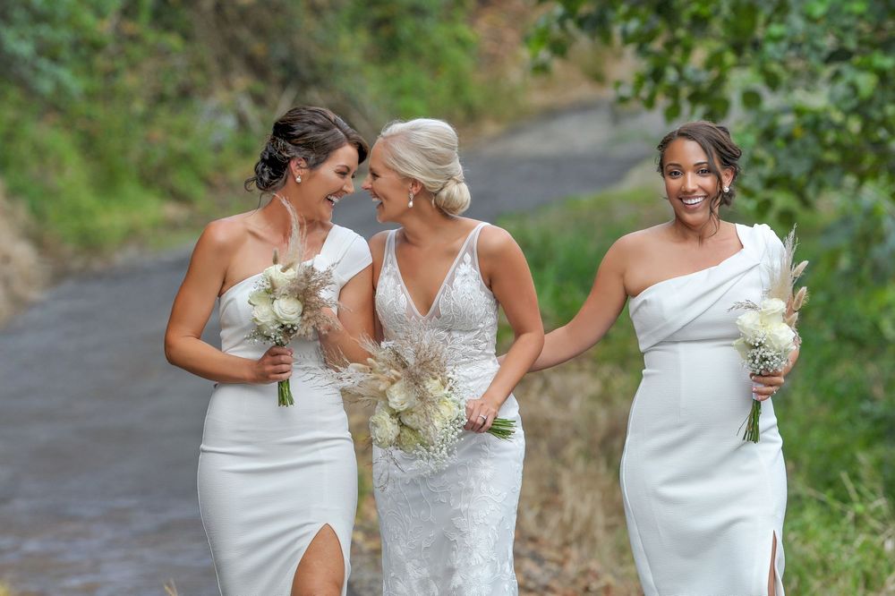 Three bridesmaids in white dresses walk together laughing along a tree-lined path with bouquets.
