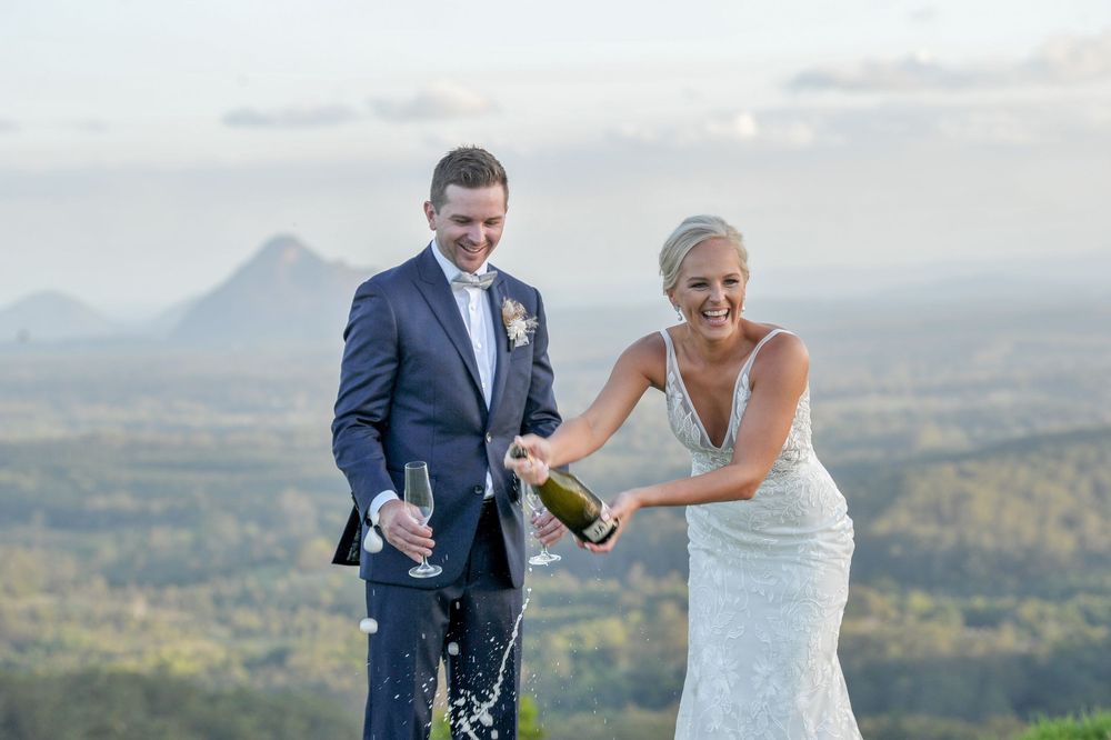 Smiling bride and groom popping champagne bottle with scenic mountain landscape in the background.