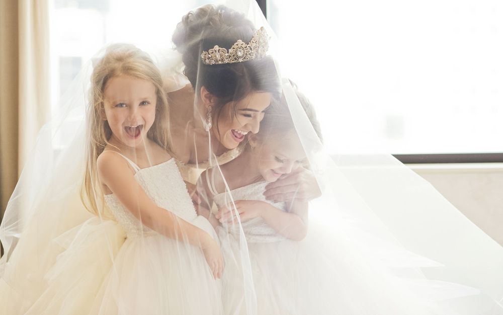 Bride in tiara and veil laughs with two young flower girls in white dresses during wedding preparations.