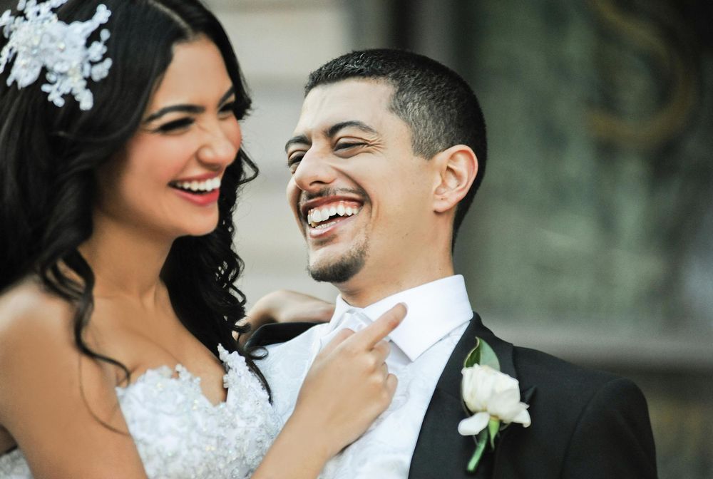 Happy bride and groom laugh together on their wedding day, groom wearing black suit with white boutonniere.