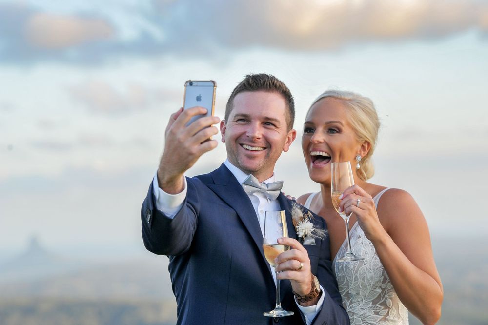 Happy wedding couple taking a selfie with champagne glasses at sunset with scenic landscape behind them.