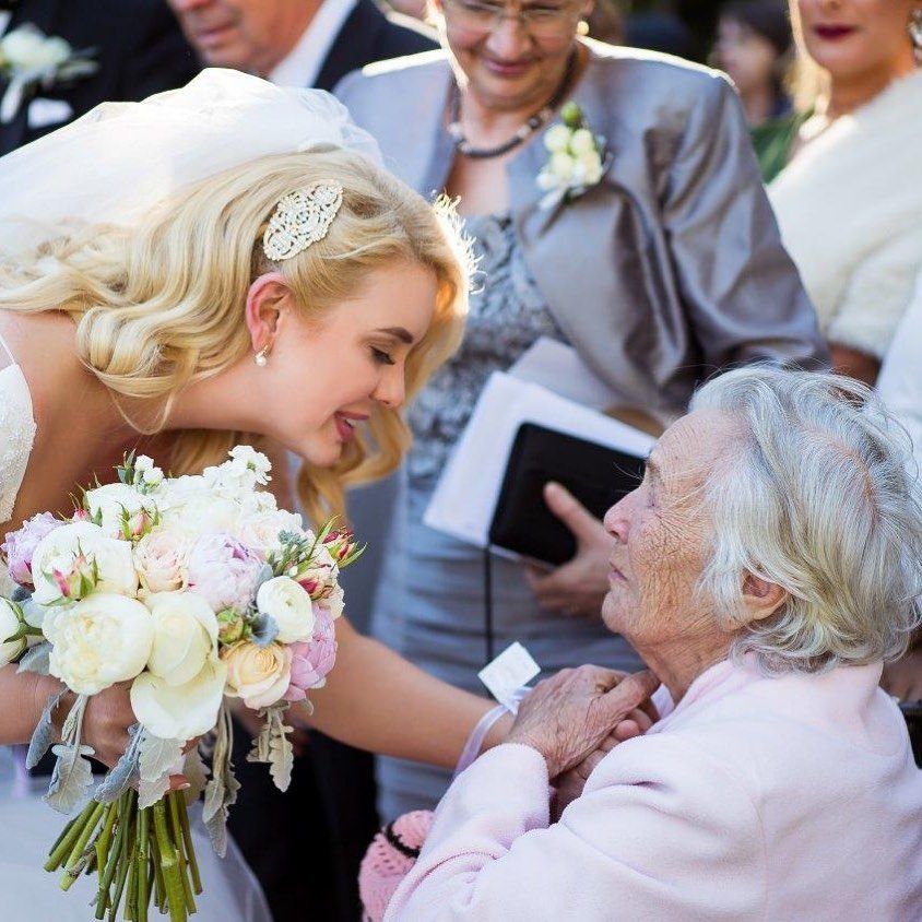 Blonde bride in lace gown with bouquet bending down to greet an elderly woman at wedding.