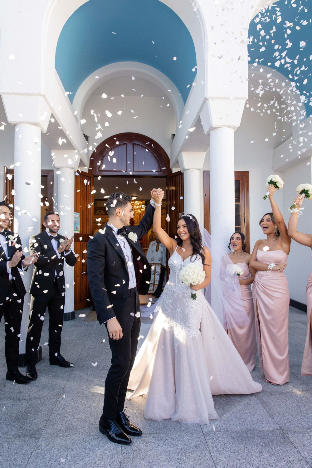 Newlyweds celebrating with confetti shower outside elegant white columned venue with wedding party.