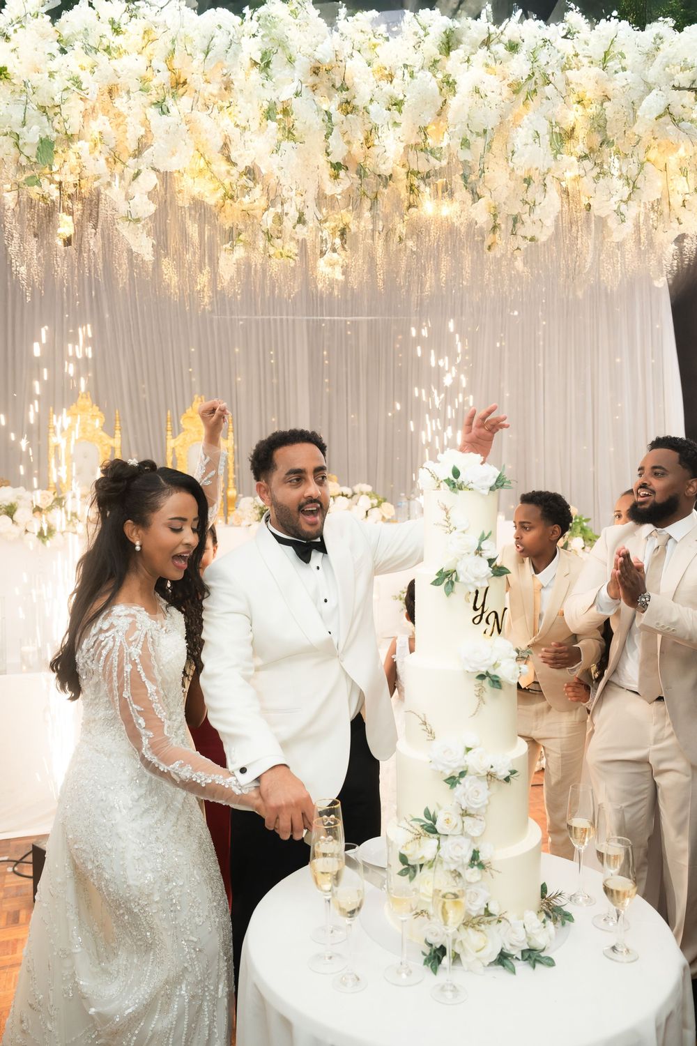 Bride and groom celebrate cutting their white floral wedding cake under glowing floral ceiling decor.