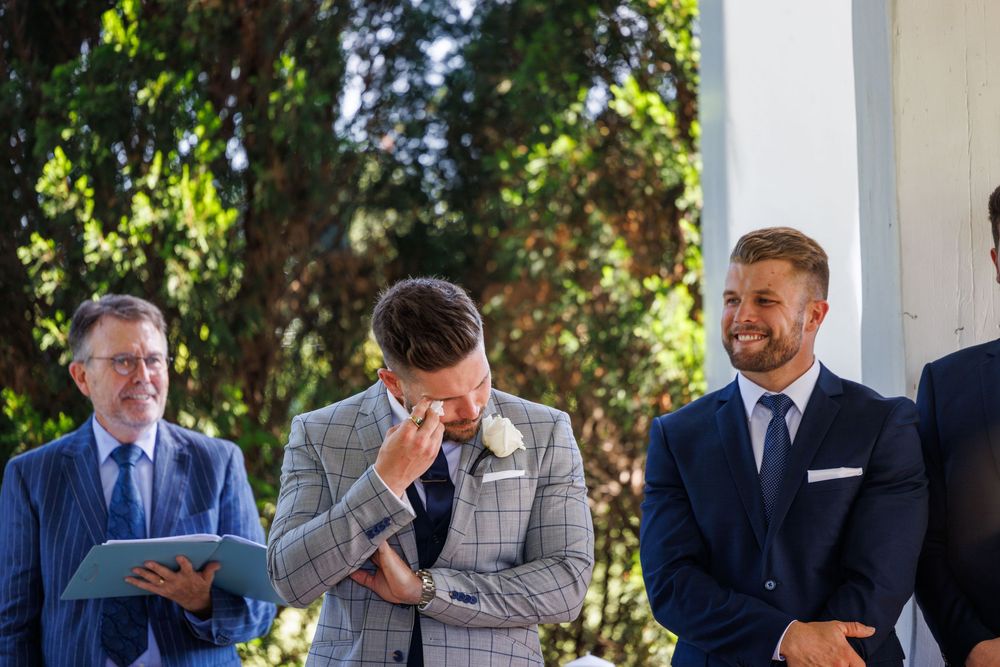 Emotional groom wiping tears at outdoor wedding ceremony with best man smiling beside him.