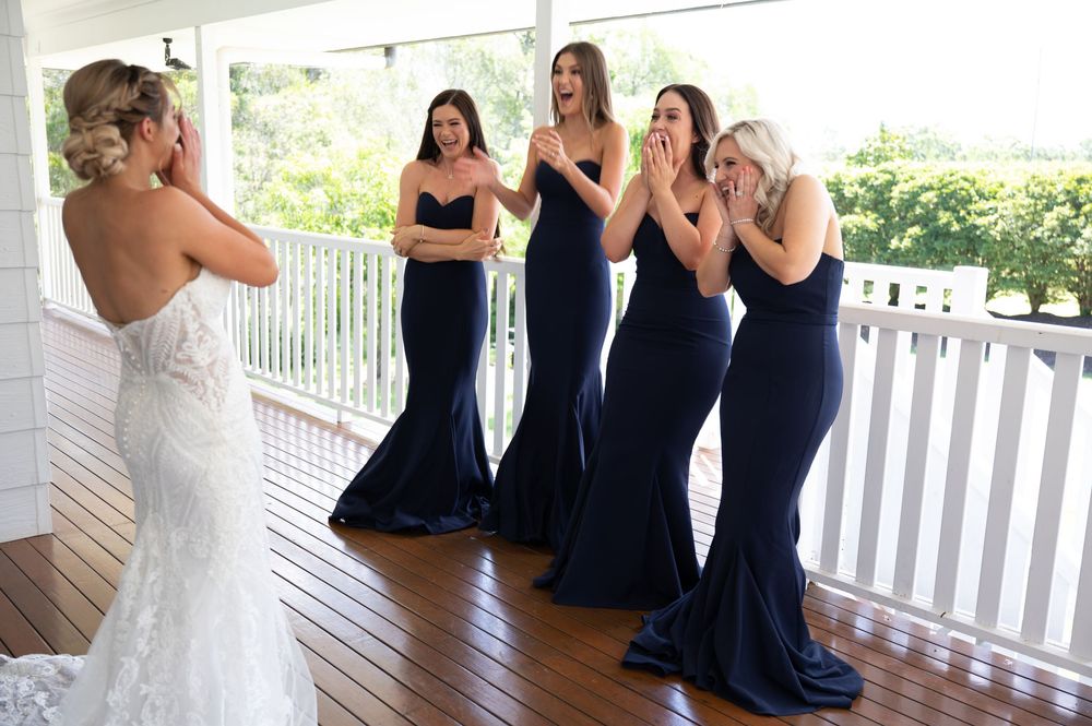 Bridesmaids in navy dresses react with joy seeing bride in white lace wedding gown on porch.