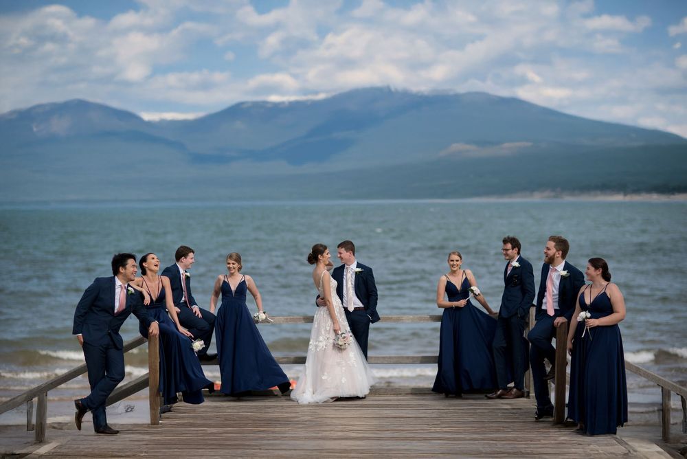 Wedding party in navy and white poses on a dock with mountain lake backdrop on a cloudy day.