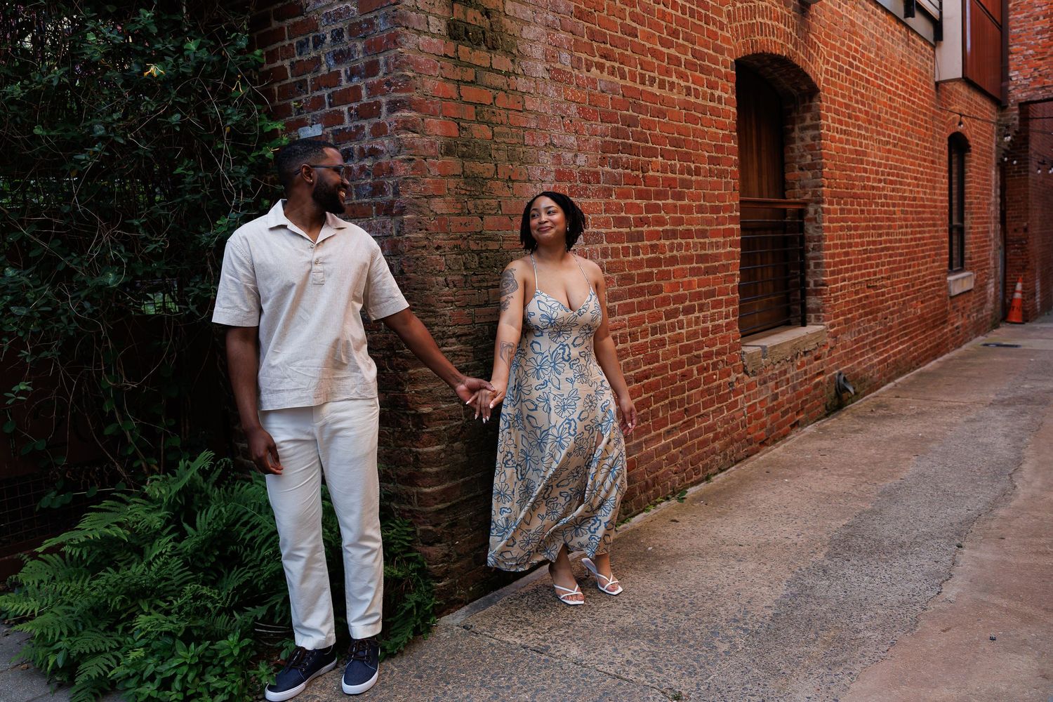 A couple walks together along a rustic brick pathway surrounded by greenery and historic architecture.