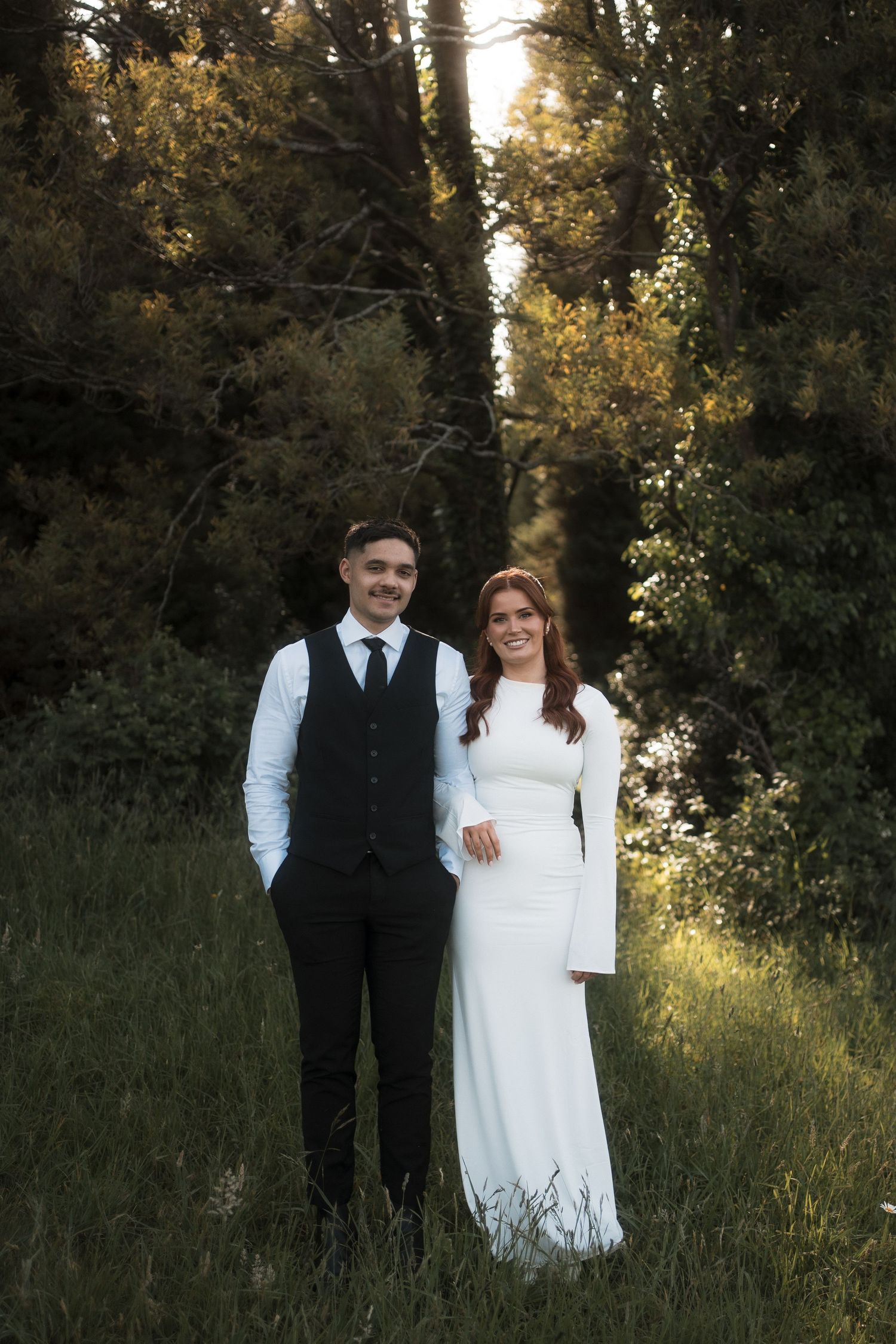 A couple in wedding attire stands together in a sunlit meadow with tall grass and trees in the background.