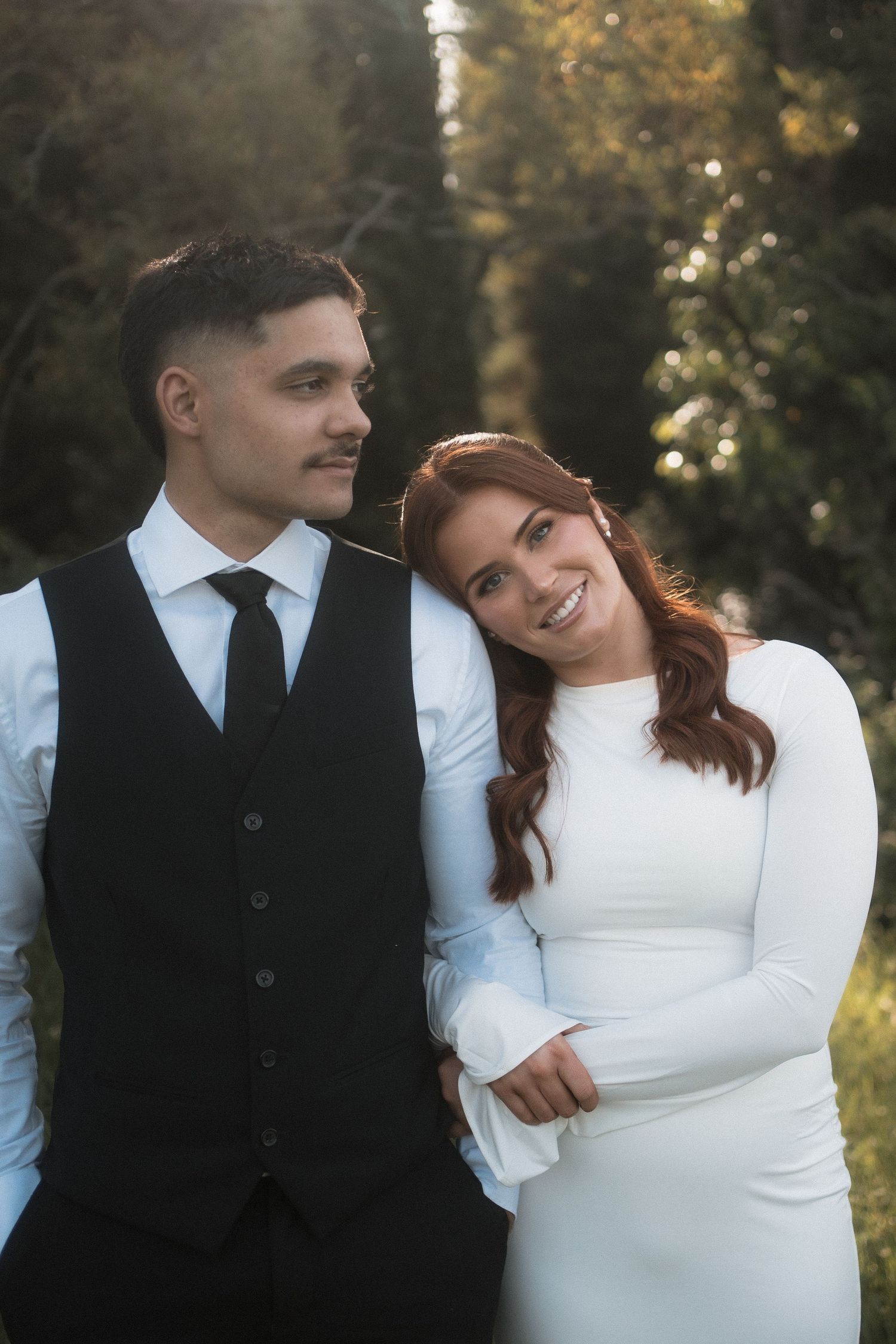 A couple in wedding attire share a joyful moment together in an outdoor setting with autumn trees in the background.