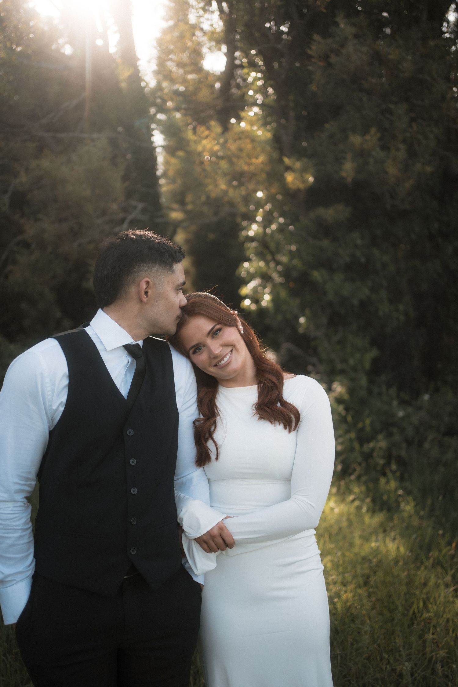 A romantic couple shares an intimate moment in a field at sunset with trees creating a natural backdrop.