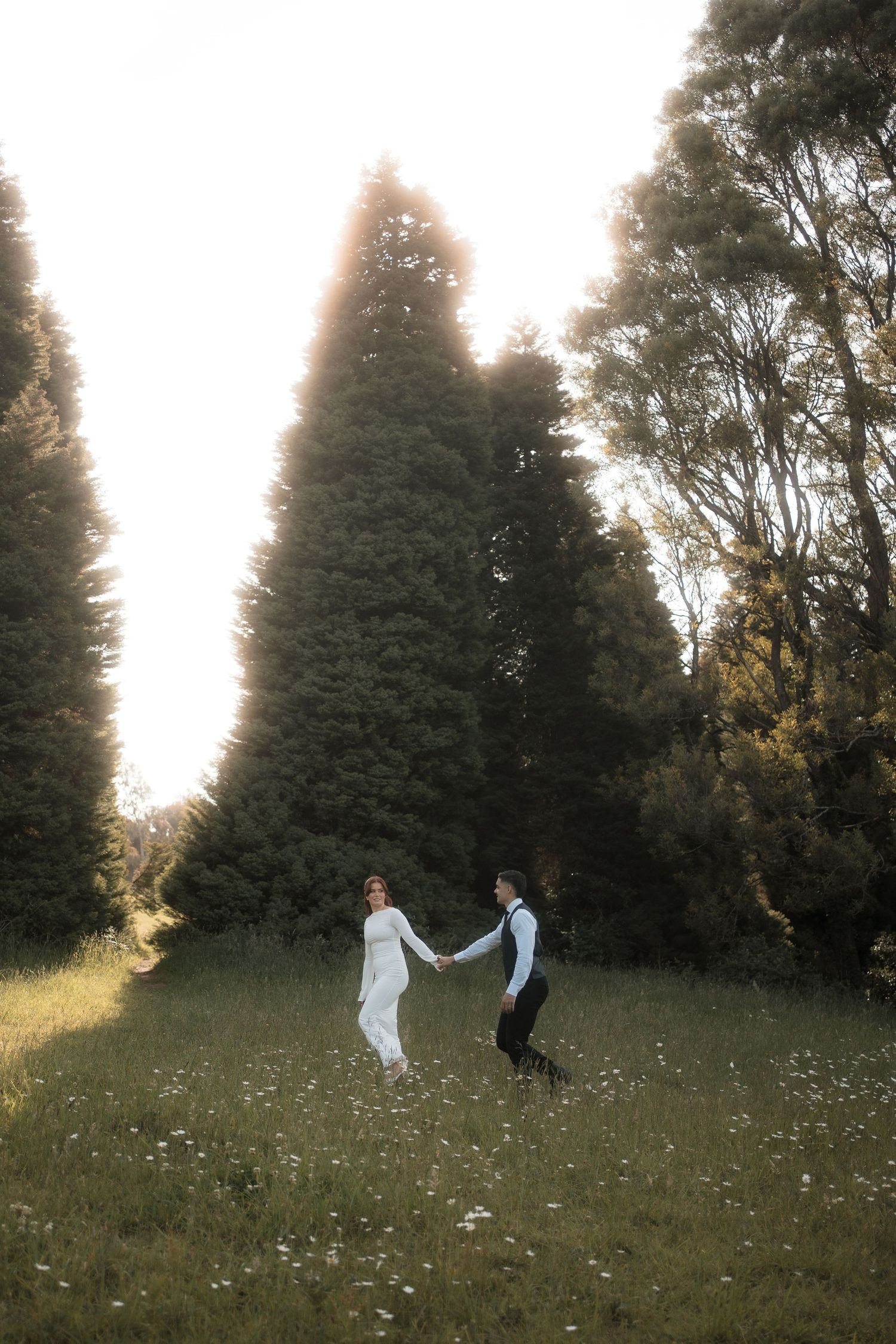 Two people dance and twirl together in a grassy meadow at sunset with evergreen trees in the background.