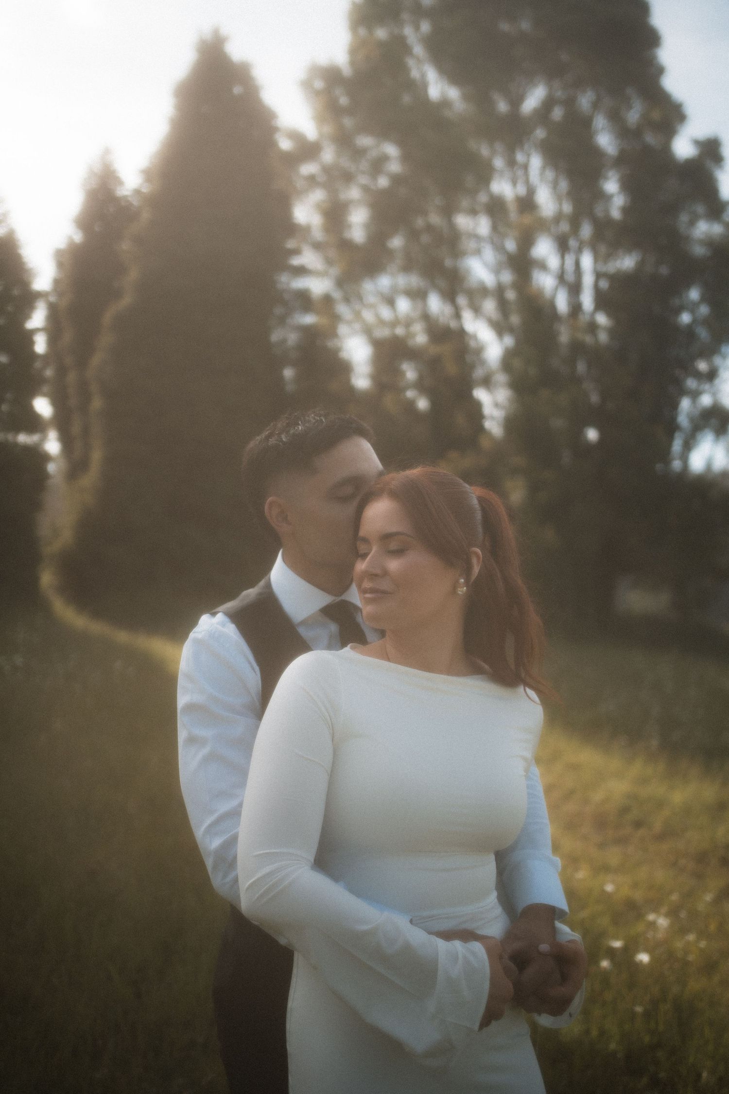 A couple embraces in a sunlit meadow with evergreen trees in the background during golden hour.